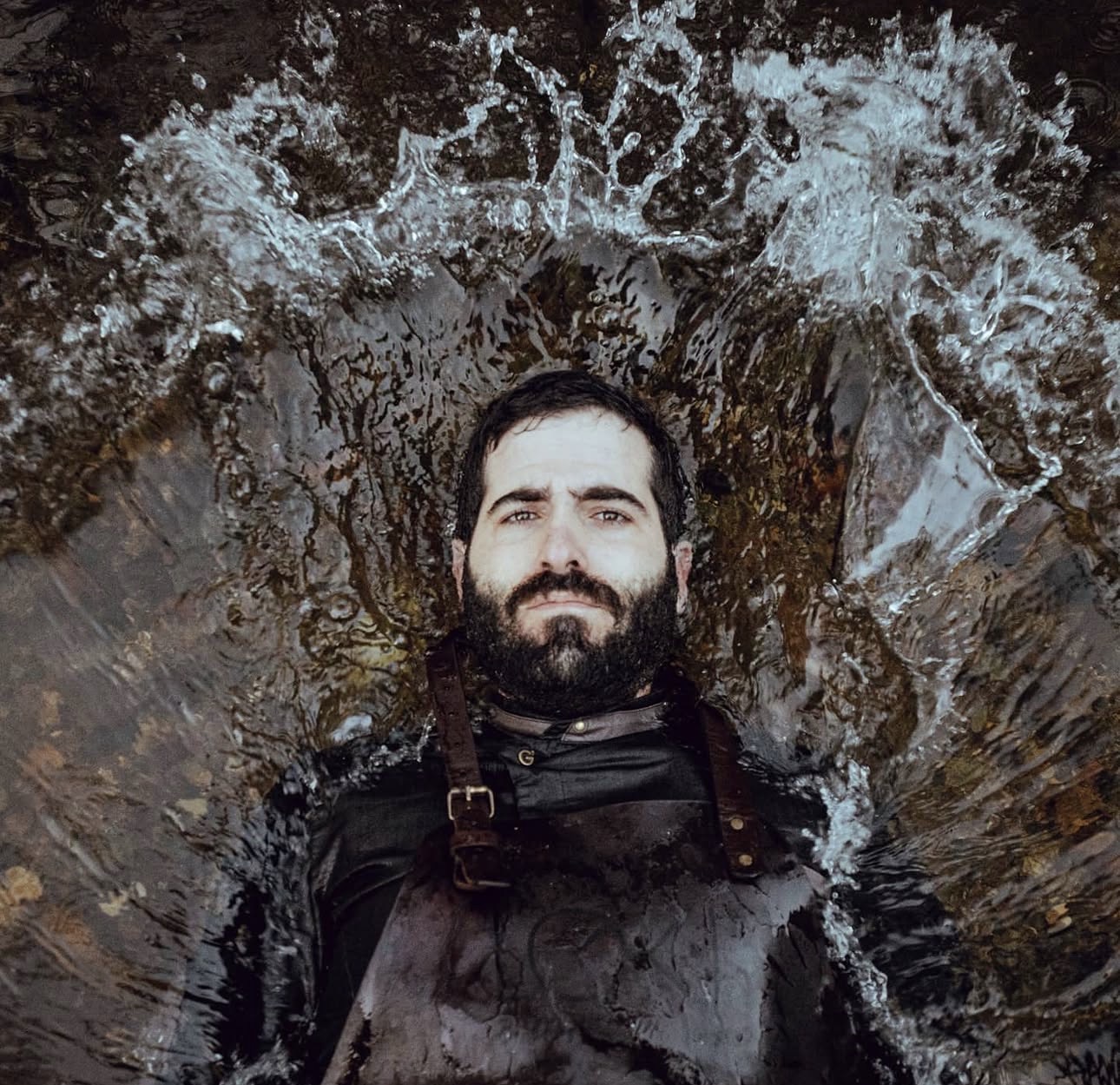 Chef João Marreiros lying in shallow water in his leather apron, water haloed around him.