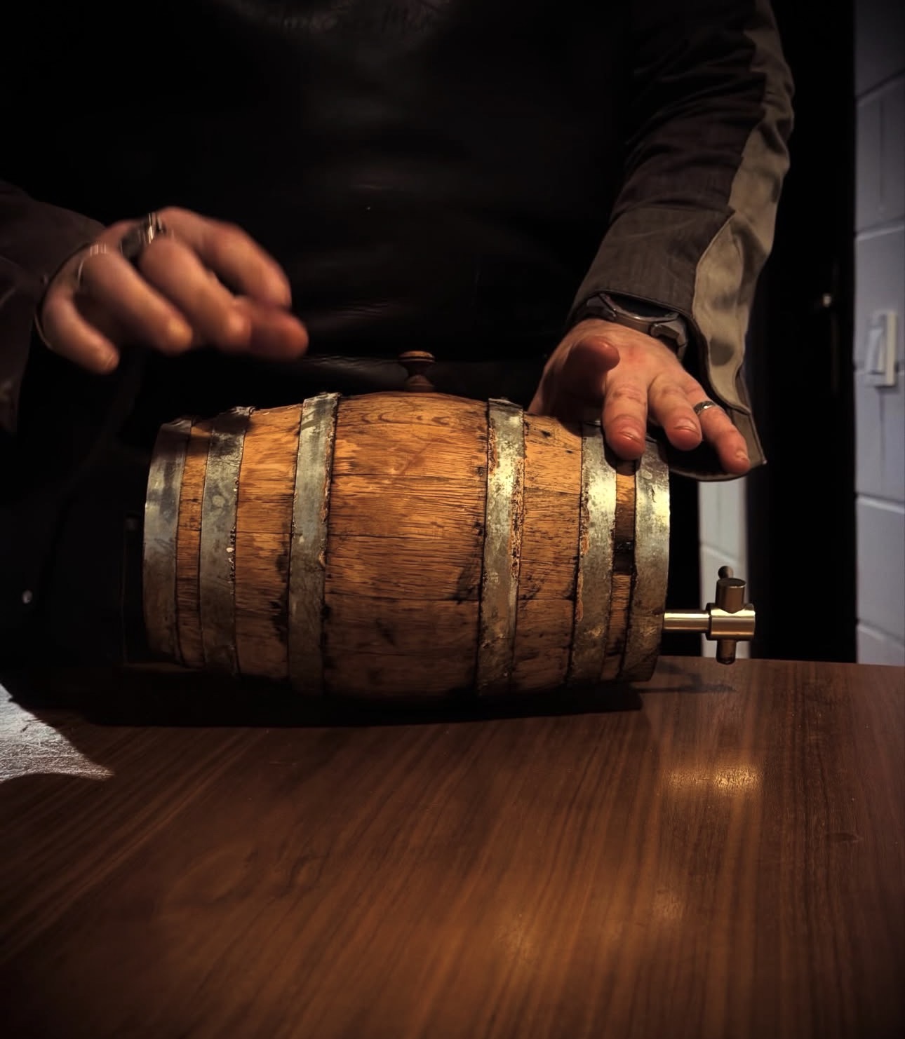 Hands rolling a small oak barrel across a dark wooden table.